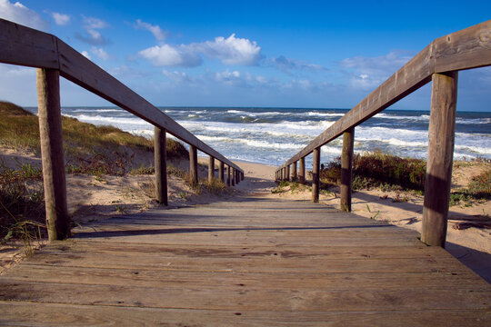 Low Angle View Of Beach Wooden Boardwalk Leading To The Rough Waves Of The Atlantic Ocean. Landscape Photography At Quiaios Beach In Portugal