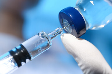 Doctor filling syringe with vaccine from vial on blue background, closeup