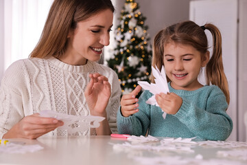 Happy mother and daughter making paper snowflakes at table near Christmas tree indoors