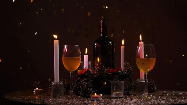 On a dark background, a New Year's still life is depicted in the form of a bottle of wine, burning candles, filled glasses and fir cones. New Year's, shiny confetti are flying onto the table.