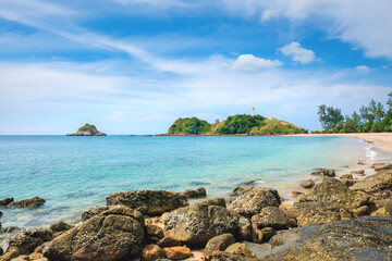 A beautiful and deserted tropical beach with yellow sand. Turquoise sea and blue sky with clouds. In the foreground are stones with oysters. A white lighthouse on a cliff. Koh Lanta, Thailand