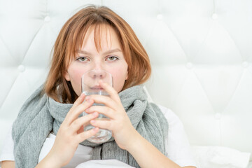 Sick girl sitting on a bed and drinking water from a glass at home. Empty space for text