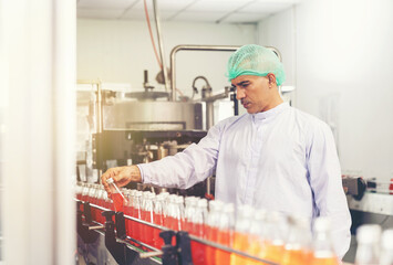 Male worker or specialist wearing hairnet and white uniform checking goods or product of Basil seed with fruit on automated conveyor belt at beverage factory. Food, beverage industry concept