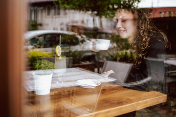 Obraz na płótnie Canvas Curly girl sitting in a cafe, drinking coffee and juice. View though the wondow