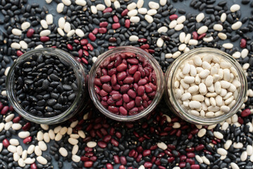 glass jar with black bean, red kidney bean and white bean. navy bean, cannelini bean, white kidney bean on  dark gray background and scattered various beans. straight line shape made from beans jar