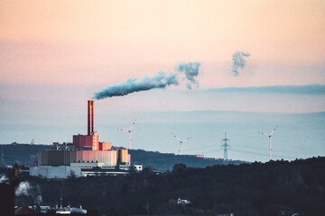 A view of a distant waste incineration plant which produces smoke which rises into the orange sky.