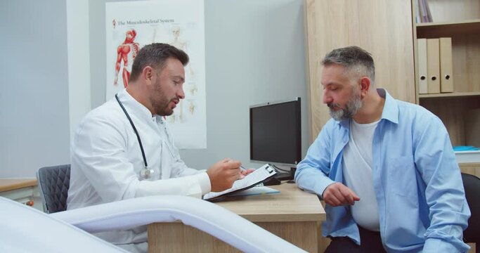 Side view. Portrait of Caucasian focused male doctor speaking and consulting middle-aged man patient about illness or surgery sitting in hospital cabinet. Handsome physician giving advice on health