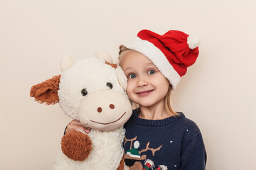 Little girl in a Christmas hat with a soft toy calf on a light background