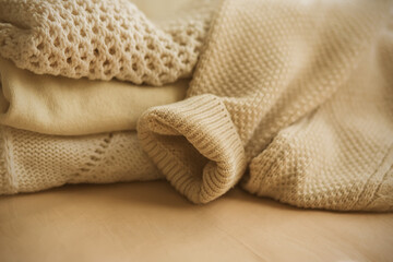 Fototapeta na wymiar Beige wool warm sweaters lie on the bed, folded in a pile, from which the sleeve of one of the sweaters has fallen. Warm clothing for cold autumn and winter.