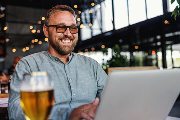 Middle aged smiling bearded man with eyeglasses sitting in his bar, using laptop and having a glass of fresh cold light beer.