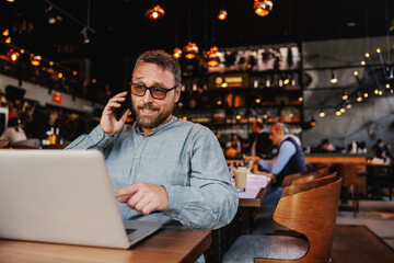 Middle aged bearded cafe owner with eyeglasses sitting in a bar, talking to financial adviser and pointing at laptop.