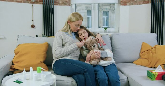 Likable Caring Adult Blond Mother Hugging Her Unhappy Sick Teen Daughter While She Measuring Temperature,holding Thermometer In The Mouth,close Up