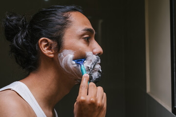 Perfil de un hombre latino que usa una navaja para afeitarse en el baño de su casa