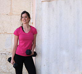 young sporty girl leaning against a wall with dumbbells and a smiling face.
