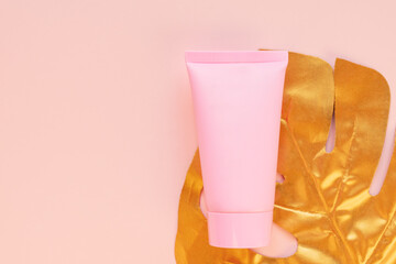 Top view of a pink tube of cream mockup with a golden monstera leaf on a pink background. Unbranded cosmetics package.