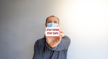 COVID-19 Pandemic symbol. White card with words 'Stay home stay safe'. A young strong man in a grey wear and medical mask. Sunshine. Beautiful white background. COVID-19 pandemic coronavirus concept.