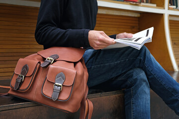 Close up man with trendy leather backpack reading magazine. Fashion accessories, stylish casual wear detail.