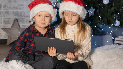 Fototapeta na wymiar children in santaclaus hats sit near a Christmas tree on a white rug with a tablet in their hands, video call with relatives on the eve of New Year's celebrations