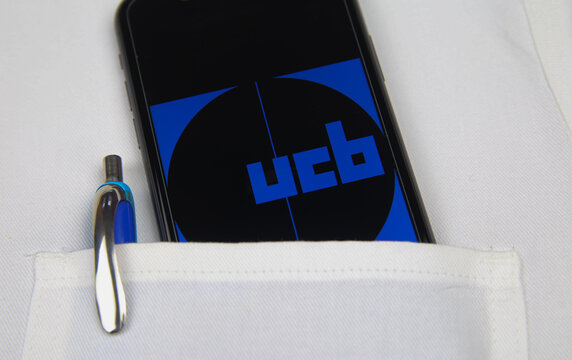 Viersen, Germany - April 9. 2020: Close up of mobile phone screen with logo lettering of UCB pharmaceutical company in pocket of white doctors coat with pencil