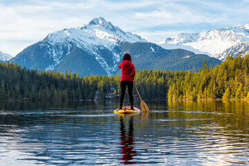 Fototapeta na wymiar Adventurous Girl Paddle Boarding on Levette Lake with famous Tantalus Mountain Range in the background. Taken in Squamish, North of Vancouver, British Columbia, Canada.