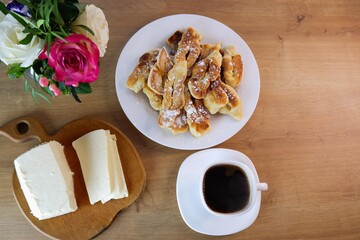 Home-baked brushwood during quarantine.  Simple food. Homemade feta cheese, a cup of coffee on a wooden table top. Concept on the background of a white brick wall and a bouquet of roses.