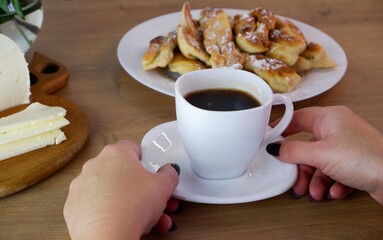 Cup of coffee in female hands, close-up. Homemade baked goods during quarantine.  Homemade feta cheese on a wooden tabletop. Concept on the background of a white brick wall and a bouquet of roses.