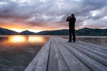 Photographing nature to spend more time outdoors and live better - A photographer standing on an old wooden pier takes a picture of a beautiful cloudy sunset with and sun setting between the mountains