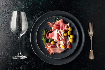 Prosciutto, green olives and rosemary on a black plate.