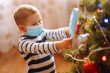 Funny little boy hangs a medical mask on the Christmas tree. Child in Sterile medical mask decoration a Christmas tree. Winter holidays  in covid-19 pandemic.