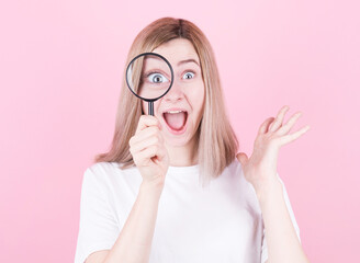 Young attractive blonde woman shocked while looking through a magnifying glass over pink background.