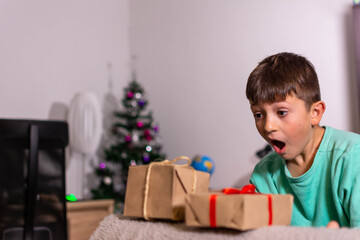 Boy sitting in a room with Christmas decor in front of a laptop with a surprised expression on his face seeing something interesting on the screen. Child spends Christmas Eve at home. Banner concept.