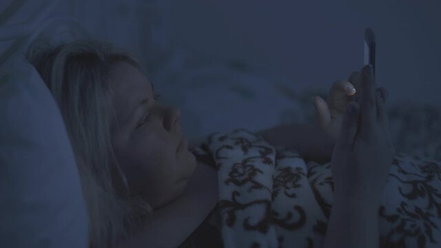 A fat woman lies in bed and checks her smart phone at night.
