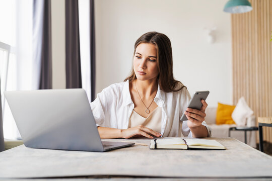 Attractive business woman sits at table in front of laptop and uses smartphone for work.