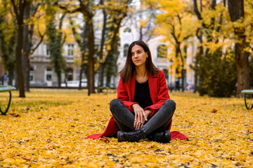 Woman in red coat sitting in the park. Around her are yellow leaves. Autumn photography.