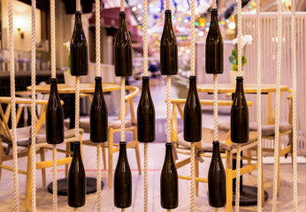 Close-up of empty wine bottles hanging from ropes. Element of creative decor of the cafe. Blurred background with festive lights.