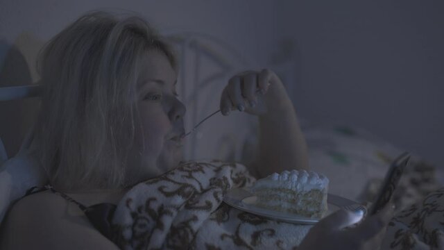 A fat woman lies in bed, eats cake and checks her smart phone at night.