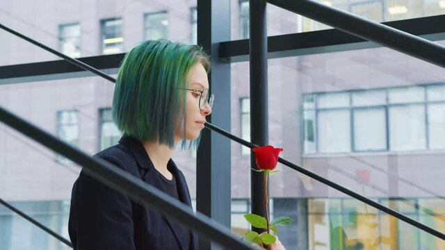 A Woman Smells A Red Rose In A Business Center. A Young Office Worker With Green Hair Sits At The Stairs Smelling A Flower. Love And Dating Site Concept