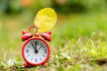 red alarm clock and yellow leaf on the grass, the concept of the end of summer