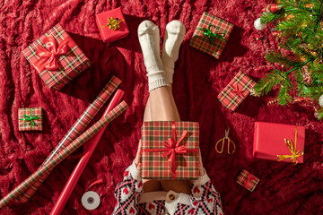 Top view of woman with christmas gift box on knees. Female wrapping gifts for family sitting on fluffy plaid near christmas tree. Flat lay.