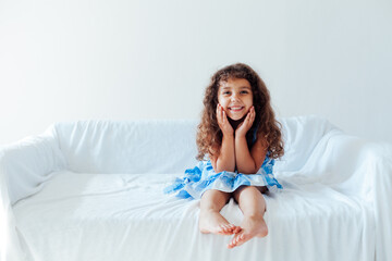 Naklejka premium Beautiful little curly girl in a blue dress fun on a white couch