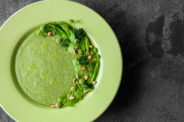 Soup of green peas, broccoli and spinach, garnished with spinach leaves, broccoli inflorescences, green peas and drops of olive oil in a green plate on a dark background