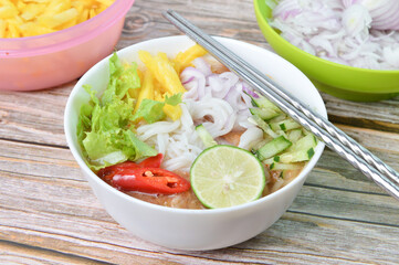 Side view of Noddle in Tangy Fish Gravy or Laksa Utara is a Special Malaysian Food Popular in Penang in a white bowl served with egg, lime, cucumber, chilies, pineapples and salads. Selective focus.
