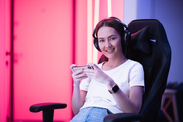 Fototapeta na wymiar Gamer girl in a big professional music headphones in white t-shirt and blue jeans holds smart phone in hands and plays game app on mobile phone, smiling and looking at camera indoors.