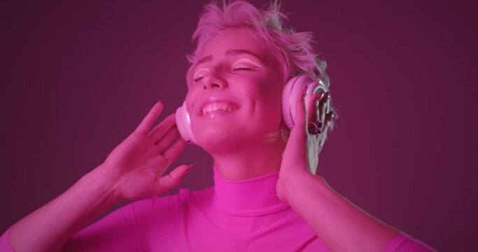 Closeup portrait of active young woman with unusual appearance, listening music in headphones, dancing, feeling excited, moving body energetically. Pink neon light background.