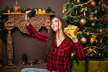 Attractive woman takes a selfie with a gift in her hands against the background of a Christmas tree.