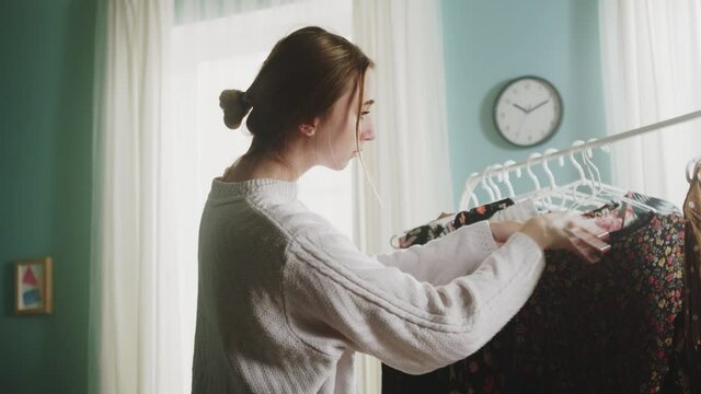 Girl in a white sweater and blue jeans stands in the middle of the room, trying on clothes and can not choose a dress