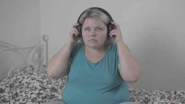 Fat woman funny sings and dances listening to music on headphones, sitting on the bed in her bedroom.