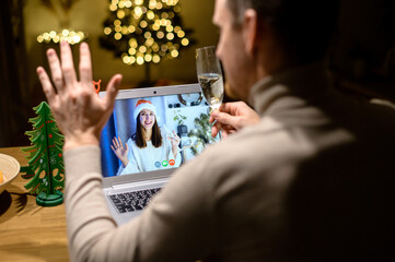 View above shoulder a guy with a glass of champagne is greetin a cheerful woman on the laptop screen. Video call, virtual celebration of Christmass eve