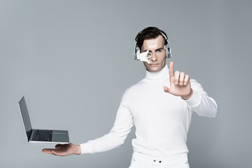 Cyborg in headphones touching something near laptop with blank screen levitating in air isolated on...
