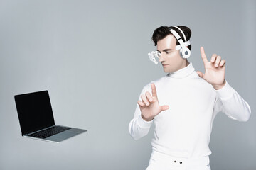 Cyborg in headphones touching something and looking at laptop with blank screen in air isolated on...